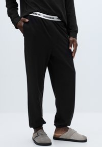 Černé teplákové kalhoty z měkké tkaniny s elastickým pasem s bílým nápisem „PULL&BEAR“. Kombinovány se šedými nazouvacími botami.