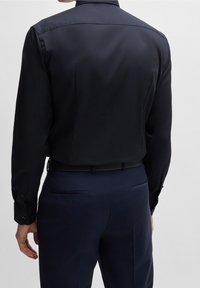 Camicia a maniche lunghe blu navy con una texture liscia, polsini con bottoni e colletto classico, indossata con pantaloni blu navy e una cintura nera.