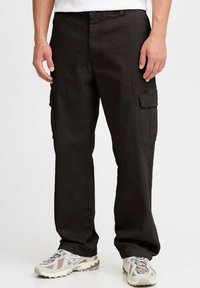 Schwarze Cargo-Hosen aus strapazierfähigem Material, mit lockerer Passform, seitlichen Taschen und dezenten Steppnähten. Getragen mit grauen Sneakers.