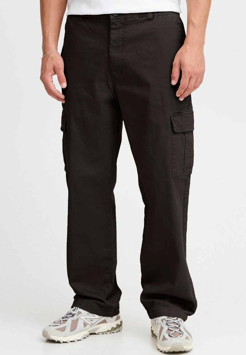 Schwarze Cargo-Hosen aus strapazierfähigem Material, mit lockerer Passform, seitlichen Taschen und dezenten Steppnähten. Getragen mit grauen Sneakers.