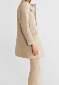 Manteau en laine beige avec col montant et double fermeture à pression, présentant une coupe droite et des poches latérales, associé à un pantalon évasé assorti.