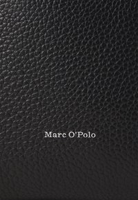 Matériau en cuir noir texturé avec un petit logo en relief "Marc O'Polo" en blanc. La surface présente un motif granuleux.