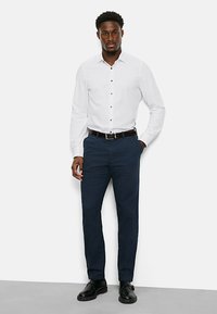 Weißes Hemd mit Knöpfen, dunkelblaue Hose, schwarzer Gürtel und schwarze Schuhe. Glattes Material, taillierter Schnitt und klassische Designelemente.