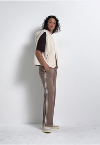 Gilet en fleece blanc cassé avec capuche, porté sur une chemise marron. Les pantalons sont de couleur marron clair avec des bandes latérales blanches. Des baskets de couleur claire complètent le look.