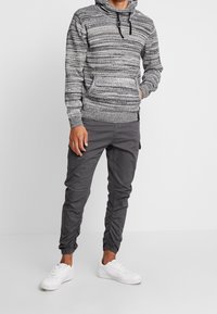 Suéter gris jaspeado con capucha y un bolsillo tipo canguro, combinado con pantalones oscuros entallados con puños elásticos y bolsillos laterales, y zapatillas blancas.