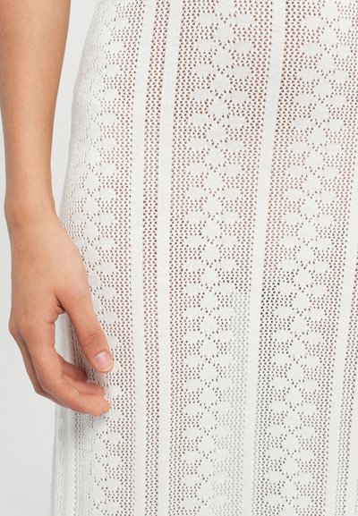 White knitted dress with a floral pattern, featuring vertical stripes and a semi-sheer texture. Hand resting on the fabric.