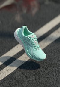 Baskets de course vert menthe avec tige en maille, semelle blanche et accents réfléchissants, flottant au-dessus d'une surface noire texturée.