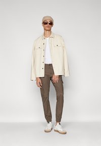Chaqueta beige oversize, camiseta blanca, pantalones cargo marrones y zapatillas blancas con acentos beige. Los accesorios incluyen gafas de sol y un gorro beige.