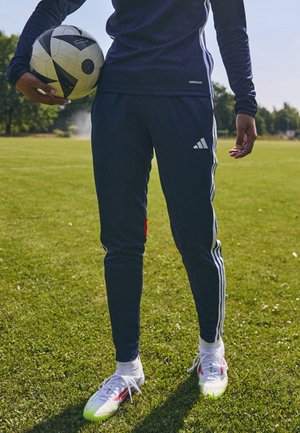 Osoba w granatowym stroju sportowym Adidas trzyma czarno-białą piłkę nożną na trawiastym boisku, w tle drzewa.