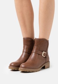 Stivali ankle in pelle marrone con zip laterale, dettaglio fibbia e suola in gomma testurizzata. Parte superiore liscia con dettagli cuciti e tacco basso.