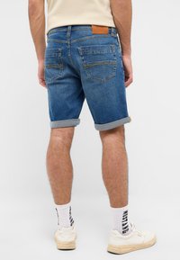 Pantalones cortos de mezclilla azul con dobladillos enrollados, dos bolsillos traseros y un parche de cuero en la cinturilla. Combinados con zapatillas blancas y calcetines.