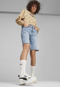 Tan sweatshirt med tryckt text, ljusblå denimshorts med fransade kanter, och kraftiga vita sneakers med gröna accenter samt ankelsockor.