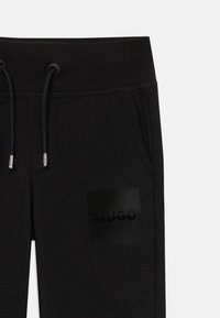 Pantalon de jogging noir avec une taille côtelée, cordon de serrage réglable, poches latérales et un patch logo « HUGO » noir mat sur la jambe.