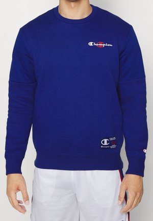 Blå sweatshirt med ribstrikkede manchetter og kant, med et lille broderet Champion-logo på brystet og en vævet etiket nederst.