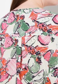 La tela de patrón floral presenta vibrantes bocetos en rosa, verde y naranja de frutas y flores sobre un fondo crema, con un diseño plisado.