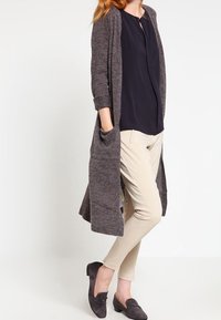 Cardigan largo de punto gris oscuro con abertura frontal, combinado con una blusa azul marino y pantalones beiges. Presenta bolsillos laterales y mocasines negros casuales.