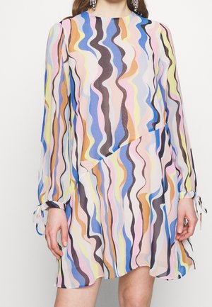 Kobieta w sukience z długim rękawem, sięgającej do kolan, z kolorowymi falistymi paskami w odcieniach niebieskiego, różowego, żółtego, brązowego i czarnego.