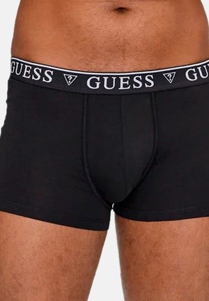 Torse masculin portant un boxer noir Guess avec une ceinture élastique de marque et un bas-ventre ainsi que le haut des cuisses visibles.