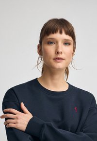 Mörkblå sweatshirt med rund halsringning, som har en liten röd logotyp på bröstet. Mjuk tyg och avslappnad passform, med långa ärmar.