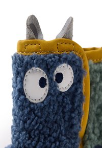 Bota azul y verde, de tela texturizada y difusa, con acentos amarillos, grandes ojos blancos circulares con un punto azul, pequeños detalles grises en forma de orejas en la parte superior.