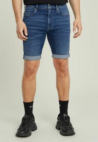 Șorturi din denim de culoare albastru închis cu mansete răsucite. Prezintă o închidere cu nasture și fermoar, buzunare frontale și un design slim fit. Se poartă adidași negri.