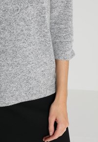 Grå stickad tröja med en mjuk textur, trekvartslånga ärmar och en avslappnad passform, tillsammans med ett svart plagg nedtill.