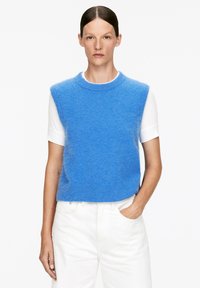 Blauer ärmelloser Strickpullover mit Rundhalsausschnitt, aus weichem Material; über einem weißen T-Shirt getragen, kombiniert mit weißen Hosen.