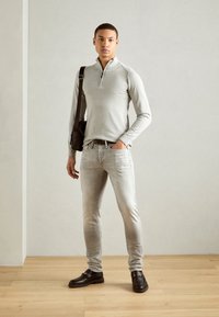 Un pull zippé gris clair, un jean gris clair décoloré, des mocassins noirs avec des détails métalliques, et un sac à bandoulière marron sur un fond neutre.