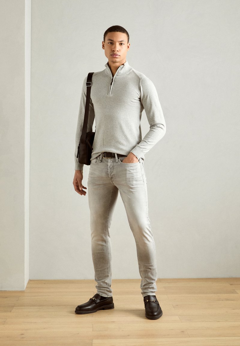 Maglione grigio chiaro con zip, jeans grigio chiaro sbiaditi, mocassini neri con dettaglio in metallo e una borsa a spalla marrone su uno sfondo neutro.