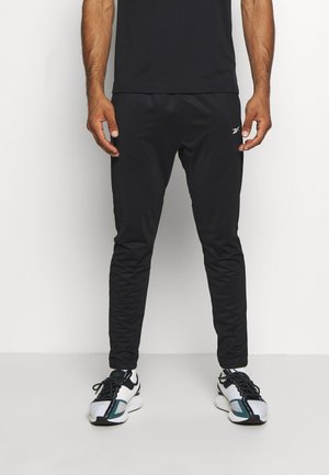 Mężczyzna ubrany w czarne zwężane spodnie Reebok i białe oraz czarne trampki stoi na białym tle.