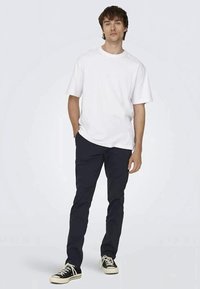 T-shirt branca de algodão com mangas curtas, combinada com calças justas azul-marinho escuras e sapatilhas pretas com solas brancas. Design simples e casual.
