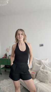 Conjunto composto por top halter preto e calções pretos, tecido liso, design justo, com bolsos, usado com brincos de argola. Mobiliário suave ao fundo.