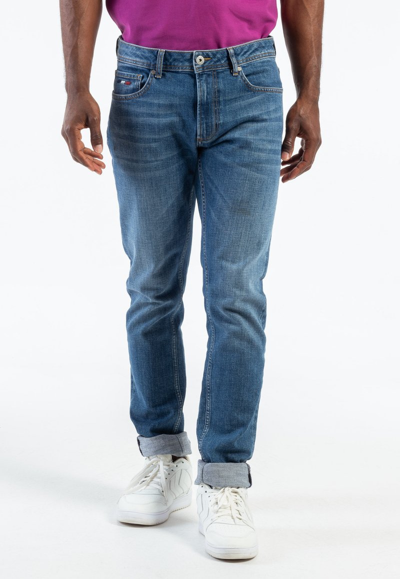 Jean en denim bleu clair avec une coupe slim, présentant un design classique à cinq poches, un léger délavage et des bas retroussés au-dessus de baskets blanches.