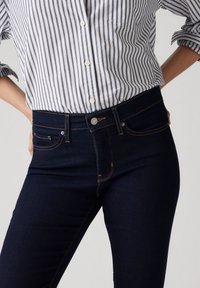 Jean en denim foncé taille haute, avec surpiqûres orange et poches avant. Associé à une chemise à rayures blanches et bleu marine à boutons.