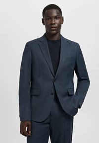 Młody mężczyzna w ciemnogranatowym garniturze i swetrze, stoi z jedną ręką w kieszeni na jednolitym jasnym tle.