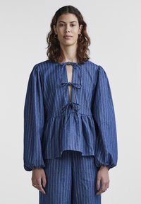 Μπλε ριγέ τζιν μπλούζα με σχέδιο peplum, ανοιχτό λαιμό και λεπτομέρειες με κορδόνια. Διαθέτει μακριά φουσκωτά μανίκια και χαλαρή γραμμή.