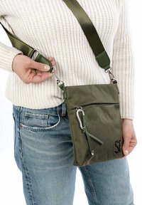 Olive green crossbody bag made of textured fabric, featuring silver hardware, an exterior zip pocket, and an adjustable strap.