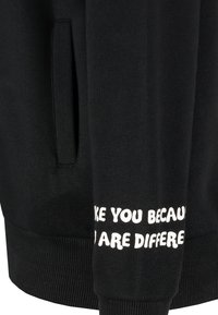 Zwarte sweatshirt met een zachte textuur, voorzien van een kangoeroe-pocket en witte tekst langs de mouw die luidt: "Ik mag je omdat je anders bent."