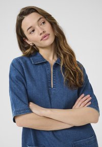 Denim tuniek in donkerblauw, met een halve knoopsluiting, korte mouwen en voorzakken. Zachte textuur met contrasterende stiksels.