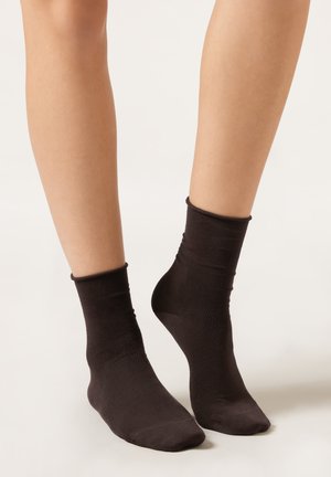 Beine mit schlichten schwarzen Knöchelsocken auf hellem Hintergrund, wobei ein Fuß leicht auf den Zehen angehoben ist.