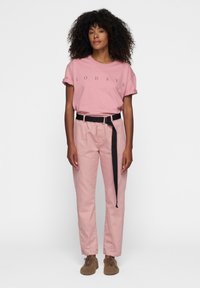 Camiseta de algodón rosa con impresión "10 DÍAS", combinada con pantalones rosas claros que tienen un cinturón negro. El modelo está de pie con las manos a los lados.