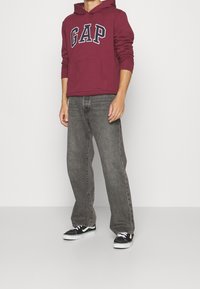 Homme portant un sweat à capuche GAP bordeaux, un jean noir délavé ample et des baskets noir et blanc, debout devant un fond uni.