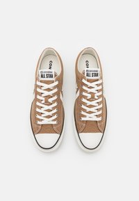 Sneaker in tela marrone con lacci bianchi, logo a stella bianca in contrasto e cappuccio in gomma. Presenta una bordatura nera sulla suola e un'etichetta con il marchio sulla linguetta.