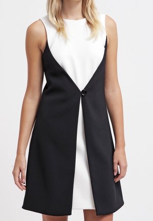 Czarna i biała sukienka bez rękawów z czarną nakładką zapinaną na jeden guzik w talii, noszona przez stojącą osobę.