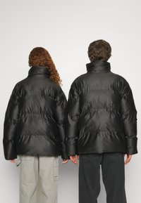 Jachete puffer negre cu guler înalt, având o textură netedă și un design matlasat. Două figuri sunt prezentate din spate, una având părul creț.