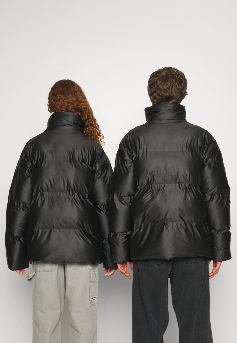 Chaquetas de plumas negras con cuellos altos, que presentan una textura suave y un diseño acolchado. Se muestran dos figuras de espaldas, una con cabello rizado.