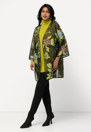Transparante zwarte kimono met bloemmotieven in groen, rood en blauw, gedragen over een felgele top en zwarte aansluitende broek, met wijde mouwen.