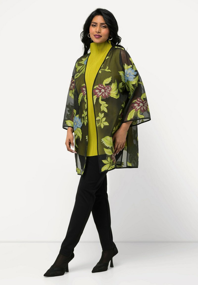 Transparante zwarte kimono met bloemmotieven in groen, rood en blauw, gedragen over een felgele top en zwarte aansluitende broek, met wijde mouwen.