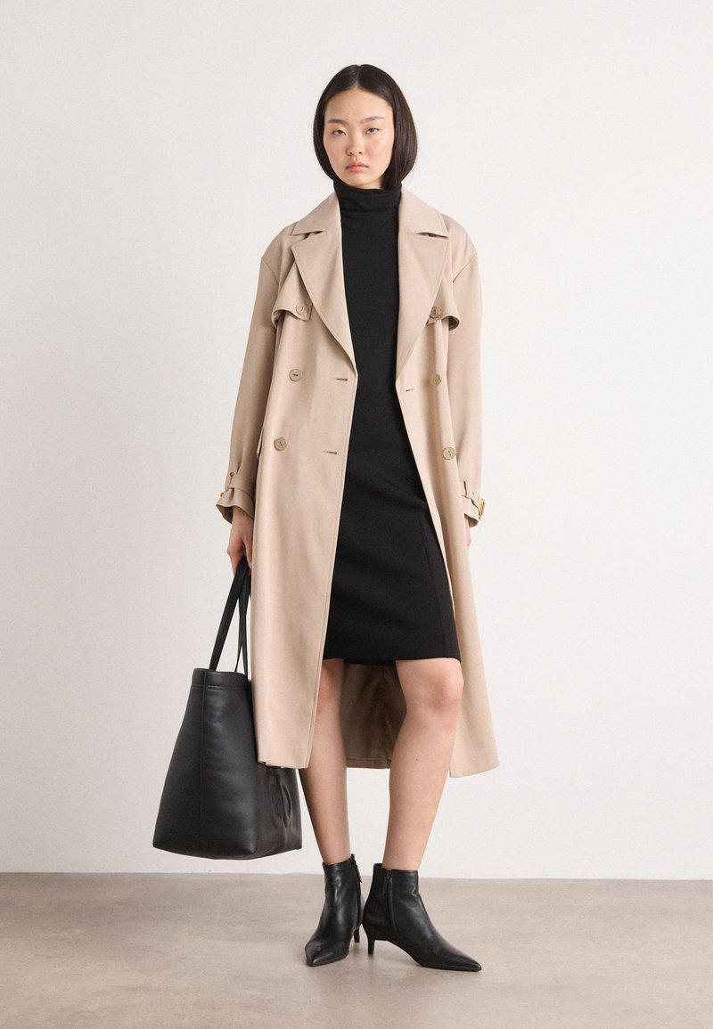 Beige Trenchcoat mit doppelter Knopfleiste, schwarzes Rollkragenkleid, schwarze Ankle Boots und eine große schwarze Tote-Tasche. Glatter Stoff und strukturierte Silhouette.