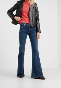 Veste courte en cuir noir portée sur un pull en maille corail, associée à un jean évasé bleu foncé et des chaussures noires, posant devant un fond uni.